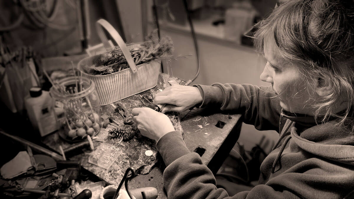 Schmuckdesignerin Ricarda Enderweit sitzt an einem Holztisch, auf dem sich getrocknete Blüten, Blätter und Werkzeuge befinden. Die Gestalterin hält ein Schmuckstück in der Hand und fräst es innen aus.
