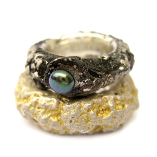 Zwei Hochzeitsringe. Ein geschwärzter Silberring mit grünlich schillernder Perle und ein breiterer Ring, der in seiner rauhen Felsstruktur zum Teil vergoldet ist.