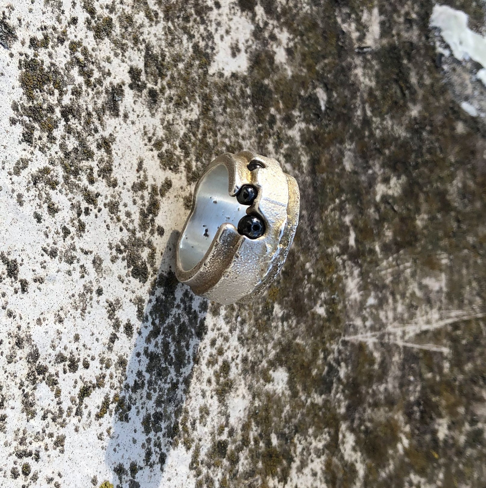 Detailaufnahme eines breiten Silberrings mit Hämatitkugeln zwischen natürlichen Flechten auf hellem Grund.