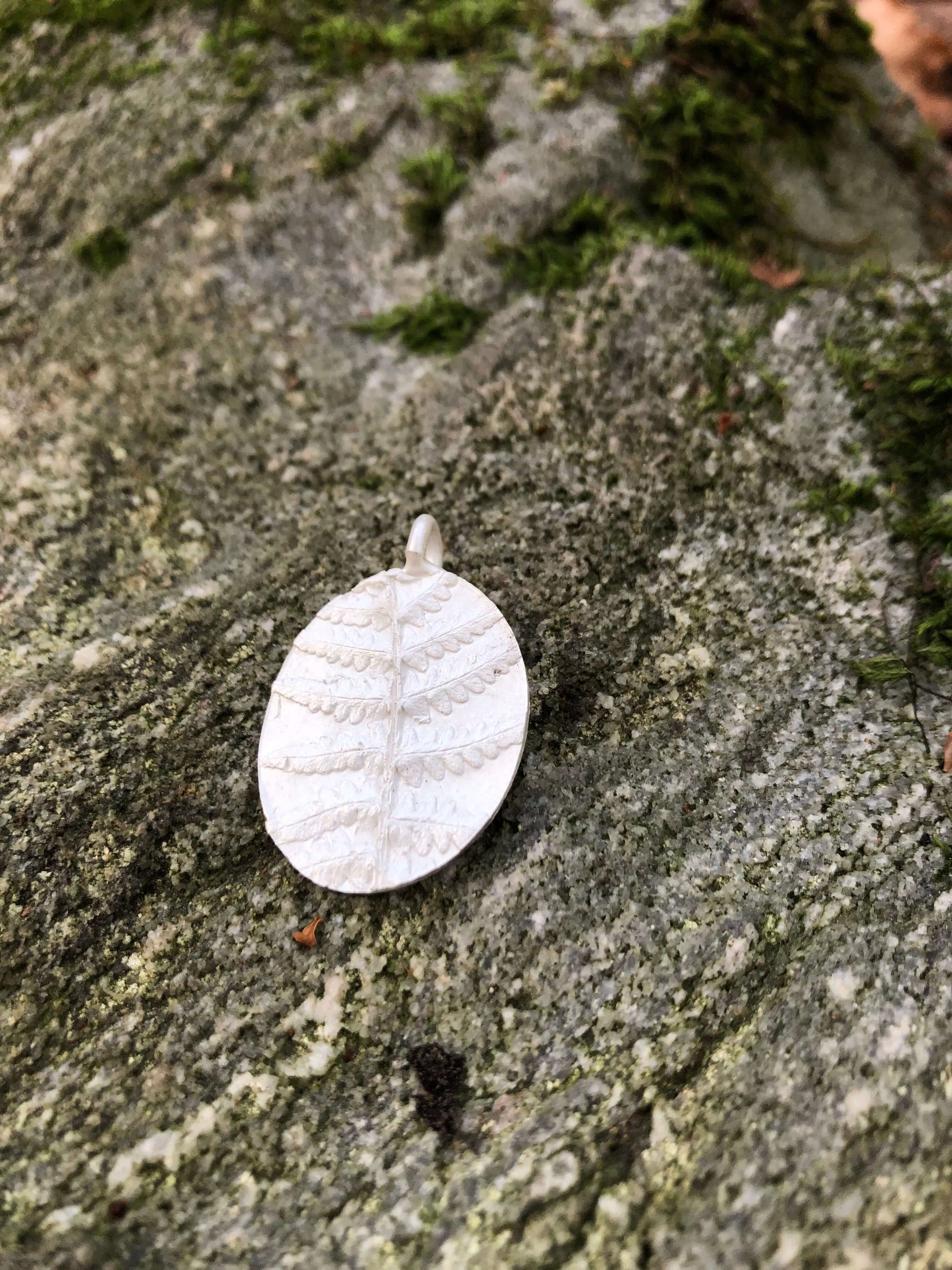 Ovaler Kettenanhänger aus Silber mit Farn auf einem moosbesetzten Felsen liegen.