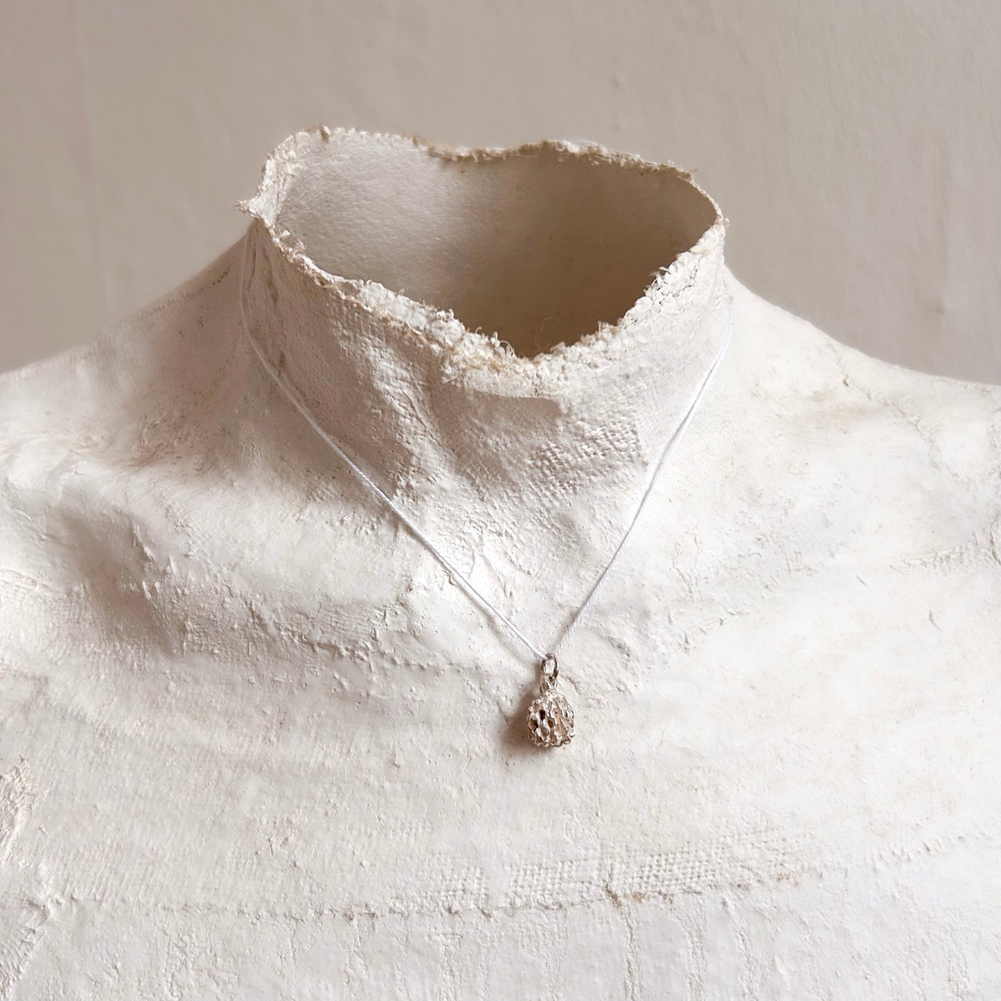 Poetische Aufnahme des Kasuarinen-Anhängers an einem Seidenfaden, getragen von einer klassischen Gipsbüste vor einer schlichten Wand.