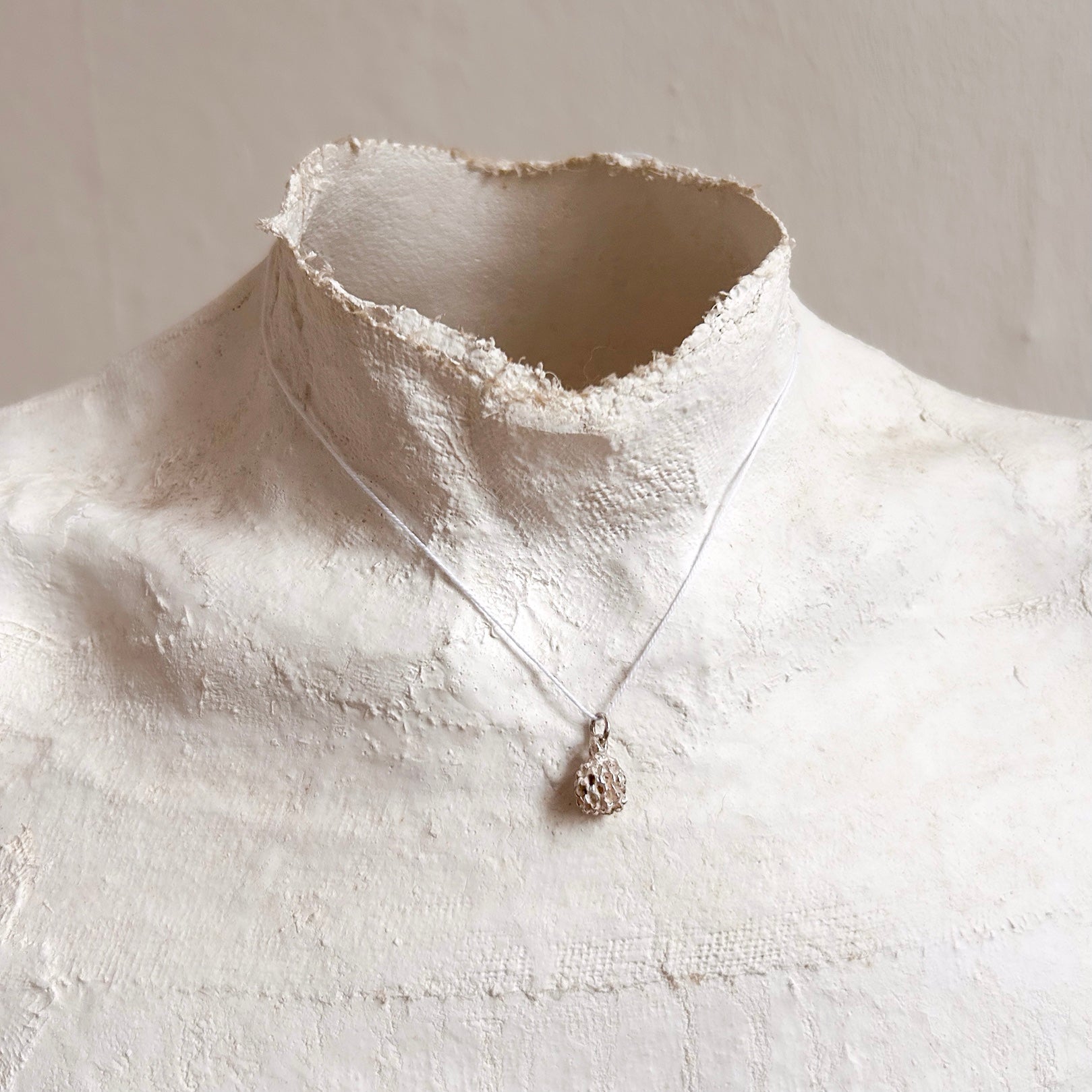 Poetische Aufnahme des Kasuarinen-Anhängers an einem Seidenfaden, getragen von einer klassischen Gipsbüste vor einer schlichten Wand.