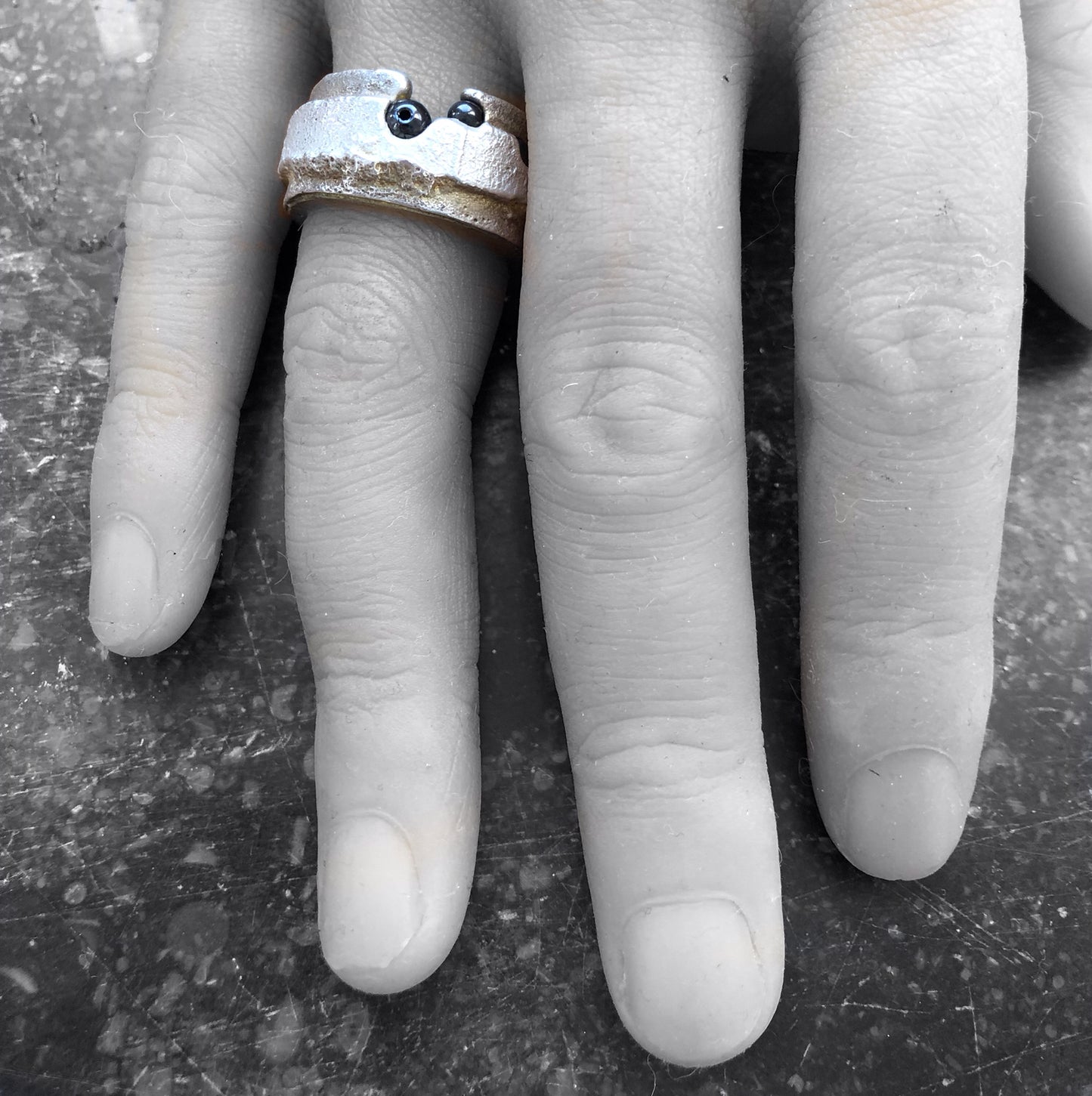 Der massive Ring am Finger, die goldigen Seiten und das Silber leuchten farbig vor einem monochromen Hintergrund.